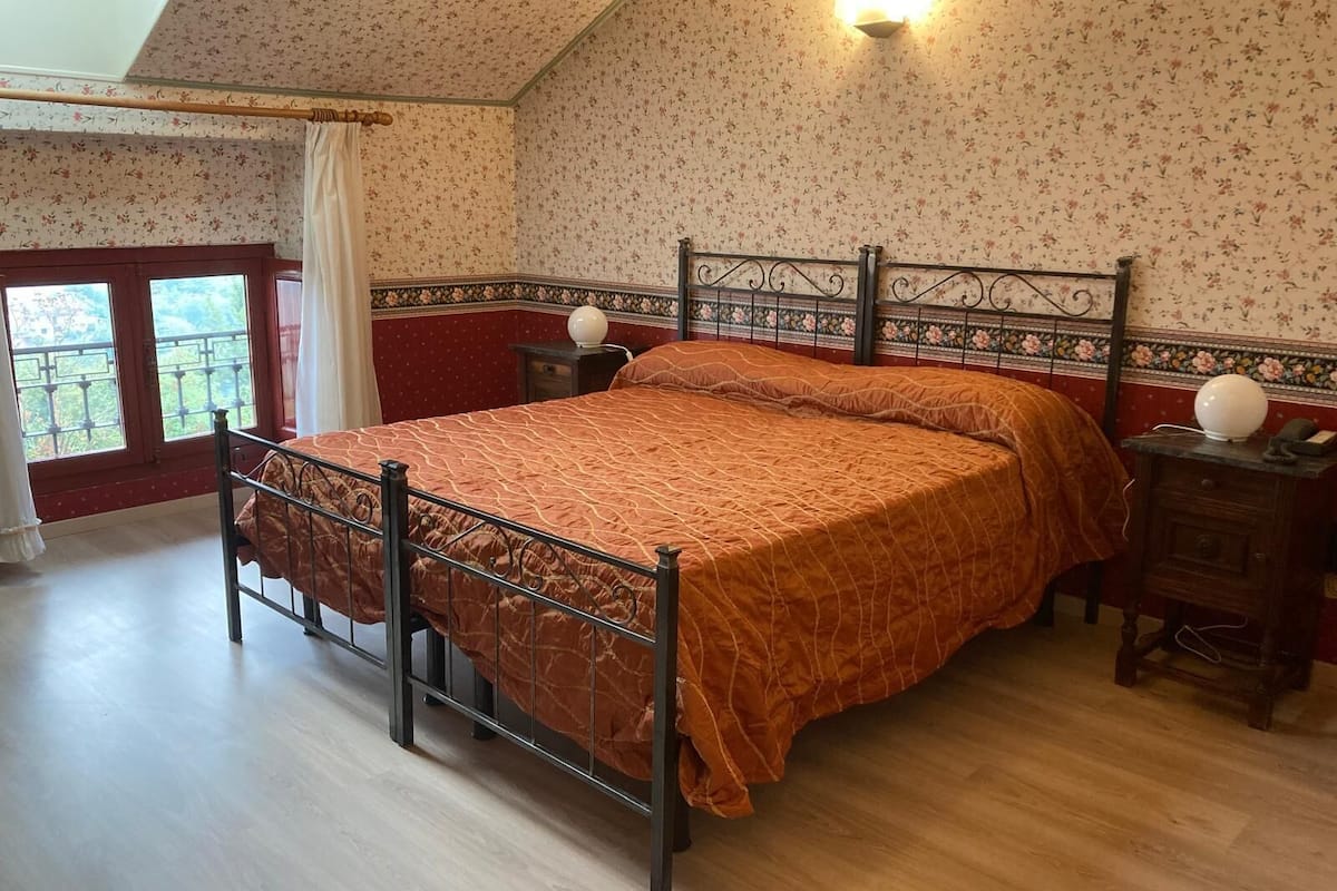 Un dormitorio con cama, mesita de noche, lámpara de pared, ventana con cortinas y vistas al balcón.