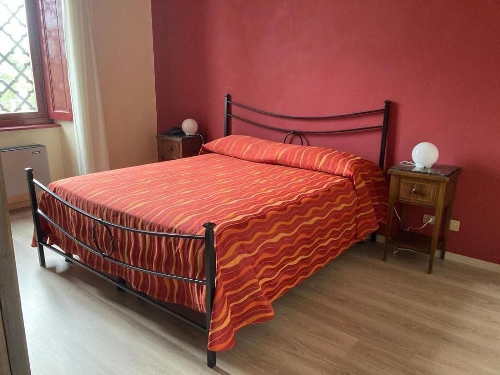 Una cama con estructura metálica y una colcha estampada roja, una mesita de noche con lámpara, y una ventana con vistas a la vegetación.