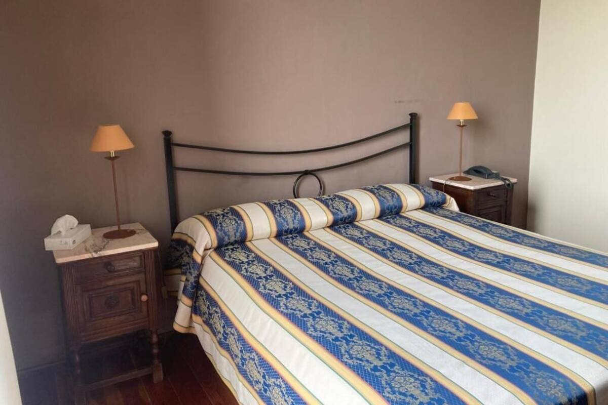 Una cama individual con colcha azul y blanca a rayas, dos mesitas de noche con lámparas y un tocador de madera con una caja de pañuelos.