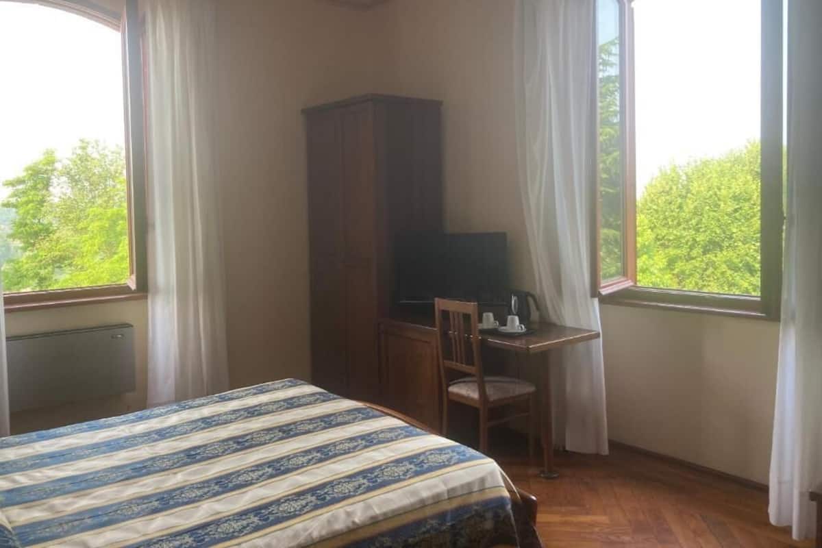 Un dormitorio con una cama, un escritorio y un televisor. Hay dos ventanas con cortinas, y se ve follaje verde afuera.