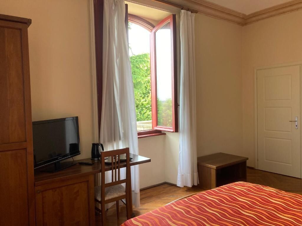 Ein Schlafzimmer mit einem Bett, einem Fernseher auf einer Holzkommode, einem Schreibtisch mit Stuhl, einem Fenster mit Vorhängen und einer Tür.