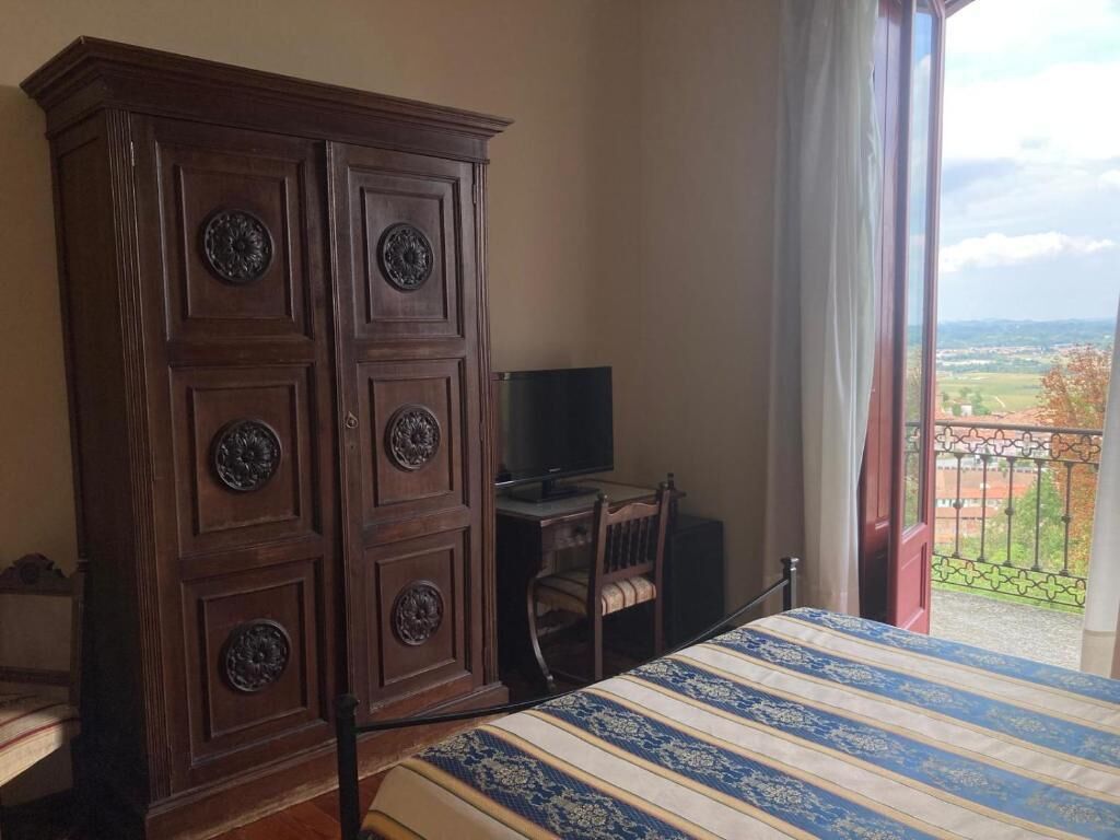 A bedroom with a large wooden wardrobe, a bed with a striped bedspread, a television on a small table, and a balcony with a view of the countryside.