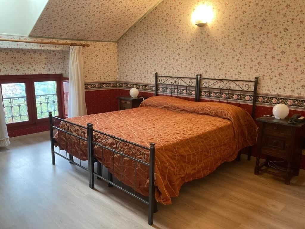 A bedroom with a bed, a nightstand, a wall-mounted lamp, and a window with a view.