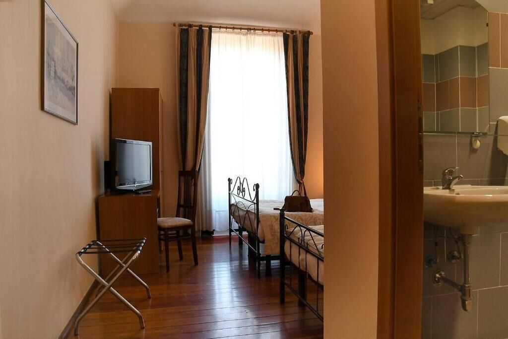 Hotellihuone, jossa on sänky, televisio, tuoli, ikkuna verhoilla ja kylpyhuone, jossa on pesuallas.