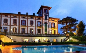A historic building with a central tower, a swimming pool, and a covered walkway with people. - hotel Villa Conte Riccardi (Rocca D'Arazzo)