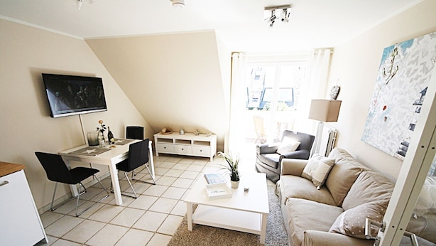 A modern living room with a white sofa, a coffee table, a TV mounted on the wall, and a dining area with a white table and chairs. - Cozy vacation apartment on the 1st floor in a quiet location in Wenningstedt (Wenningstedt)