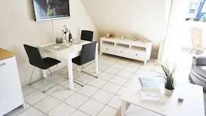 A modern, well-lit room with a dining table, chairs, a white cabinet, and a television mounted on the wall. - Cozy vacation apartment on the 1st floor in a quiet location in Wenningstedt (Wenningstedt)