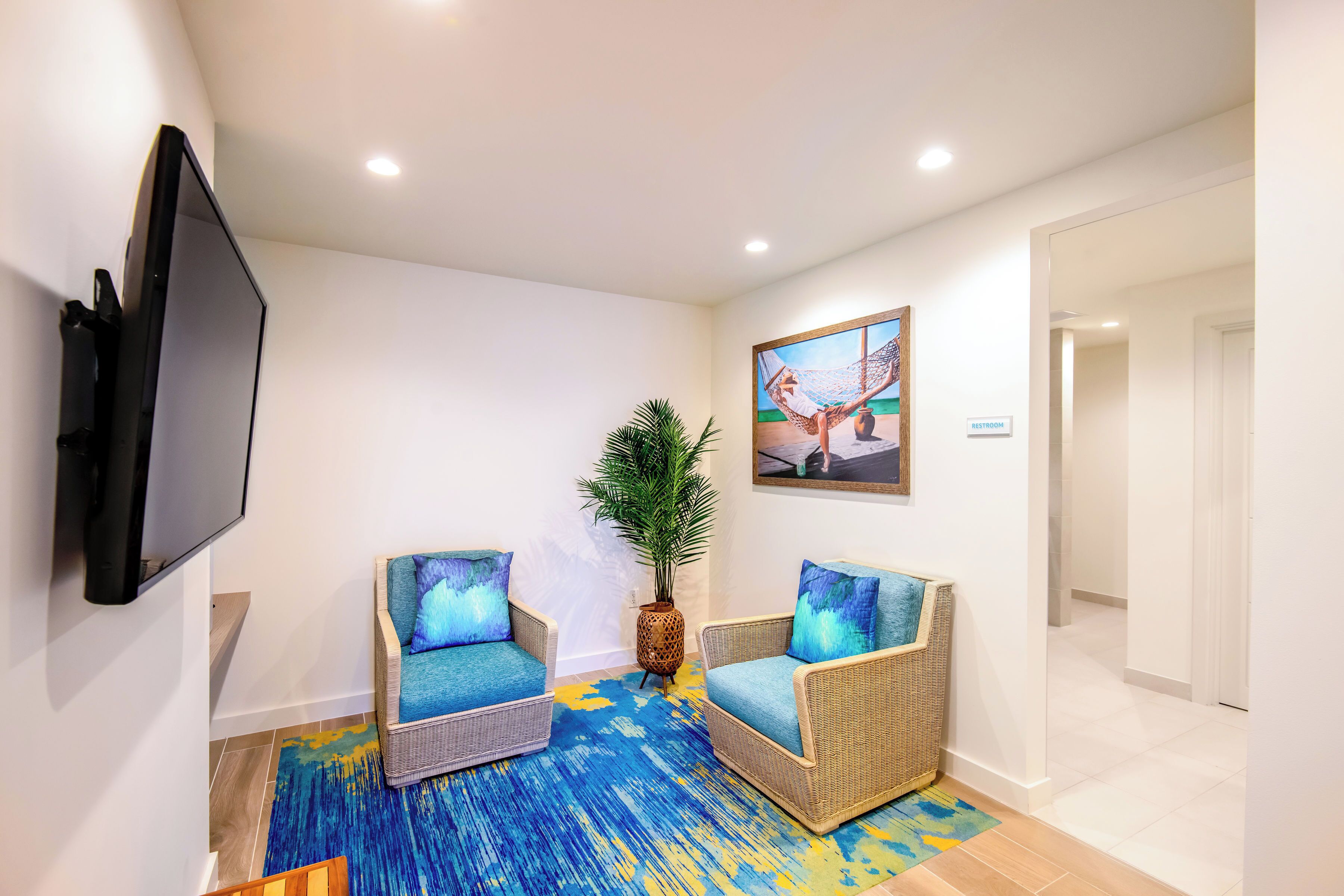 A room with two wicker chairs with blue cushions, a large flat-screen TV mounted on the wall, a painting above the TV, and a potted plant between the chairs.