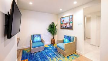 A room with two wicker chairs with blue cushions, a large flat-screen TV mounted on the wall, a painting above the TV, and a potted plant between the chairs.
