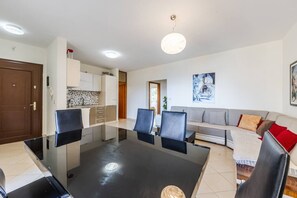 Een moderne woonkamer met een glazen eettafel, een grijze bank en een keuken op de achtergrond.