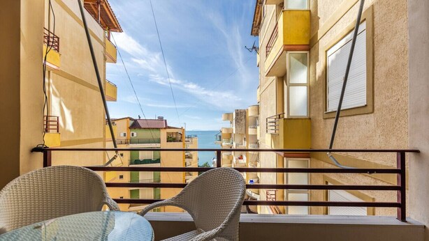 Ein Balkon mit Korbstühlen und einem Glastisch, mit Blick auf eine Küstenlandschaft mit Gebäuden und dem Meer.