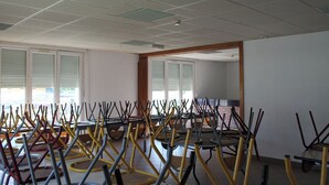 Una habitación con varias mesas y sillas dispuestas en círculo, ventanas amplias con persianas y un techo con iluminación empotrada.