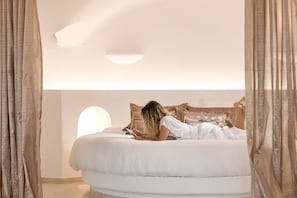 Un grand lit avec une tête de lit blanche et de nombreux oreillers, une personne allongée sur le lit en train de lire un livre, et des rideaux légers aux fenêtres.
