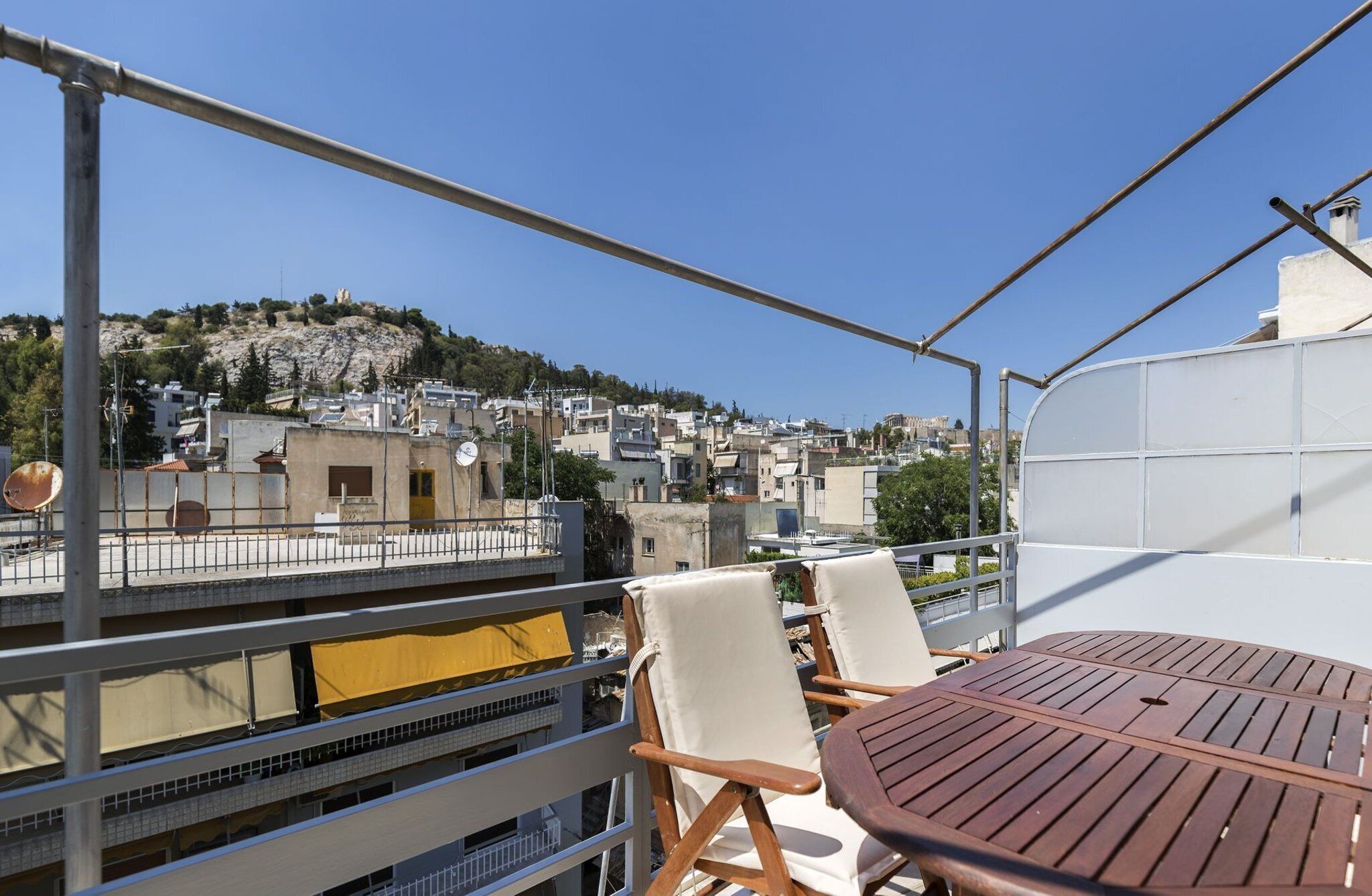 A balcony with a table and chairs, overlooking a cityscape with buildings and a hill.