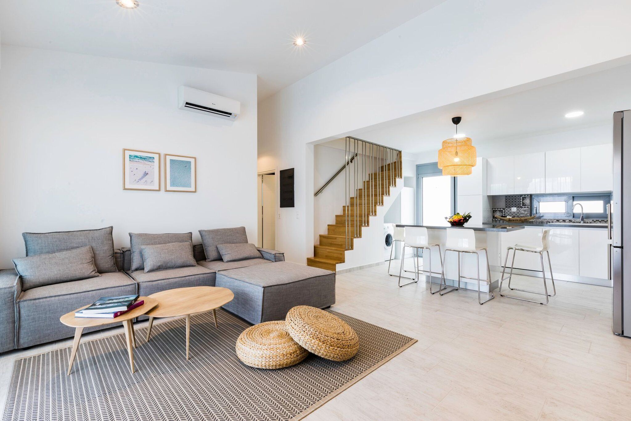 A modern living room with a grey sofa, a wooden coffee table, and a patterned rug. A staircase leads to a kitchen area with white cabinets and a stainless steel refrigerator.