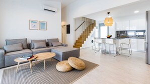 Un salon moderne avec un canapé gris, une table basse en bois et un tapis à motifs. Un escalier mène à une cuisine équipée de meubles blancs et d’un réfrigérateur en acier inoxydable.