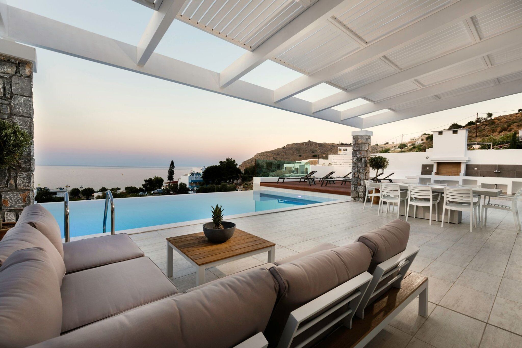 Ein moderner Poolbereich im Freien mit Blick auf das Meer, einer Steinmauer und einer Sitzecke mit Tisch und Kissen.