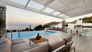 Area piscina moderna all'aperto con vista sul mare, una parete in pietra e un salotto con tavolo e cuscini.