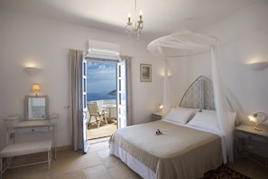 Ein Schlafzimmer mit Bett, Nachttisch, Lampe und Blick auf das Meer durch eine offene Tür.