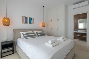 Ein ordentlich eingerichtetes Schlafzimmer mit einem großen Bett, zwei Nachttischlampen, einem Nachttisch und einem Badezimmer mit Waschbecken und Spiegel.