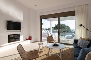 Ein modernes Wohnzimmer mit Flachbildfernseher, Korbmöbeln, einem runden Couchtisch und Blick auf einen Außenpool.