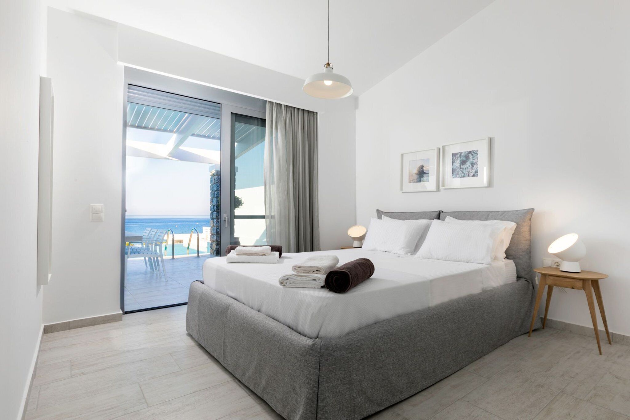 Een moderne slaapkamer met een groot bed, uitzicht op de zee door een schuifdeur, en een klein nachtkastje met een lamp.