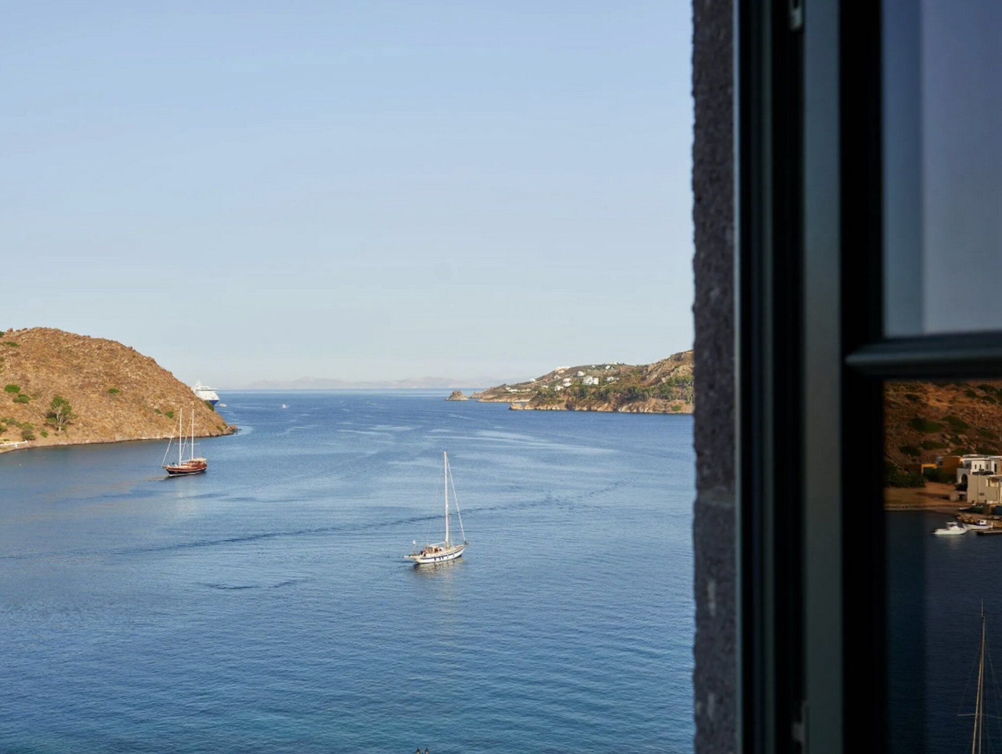 Vista costera con un velero y un barco más grande a lo lejos, una costa rocosa y edificios a la derecha.