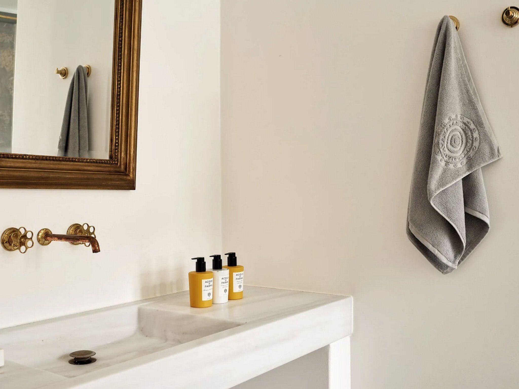 A bathroom with a white sink, a mirror with a gold frame, a towel hanging on the wall, and three bottles of toiletries on the counter.