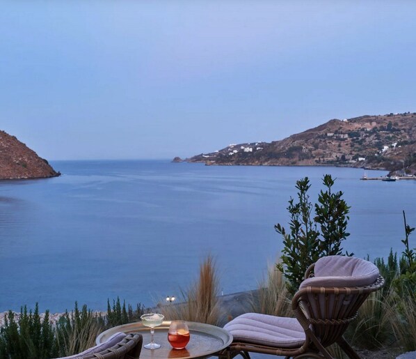 A seaside terrace with wicker furniture, a table with a drink and a candle, and a view of the sea and distant hills.