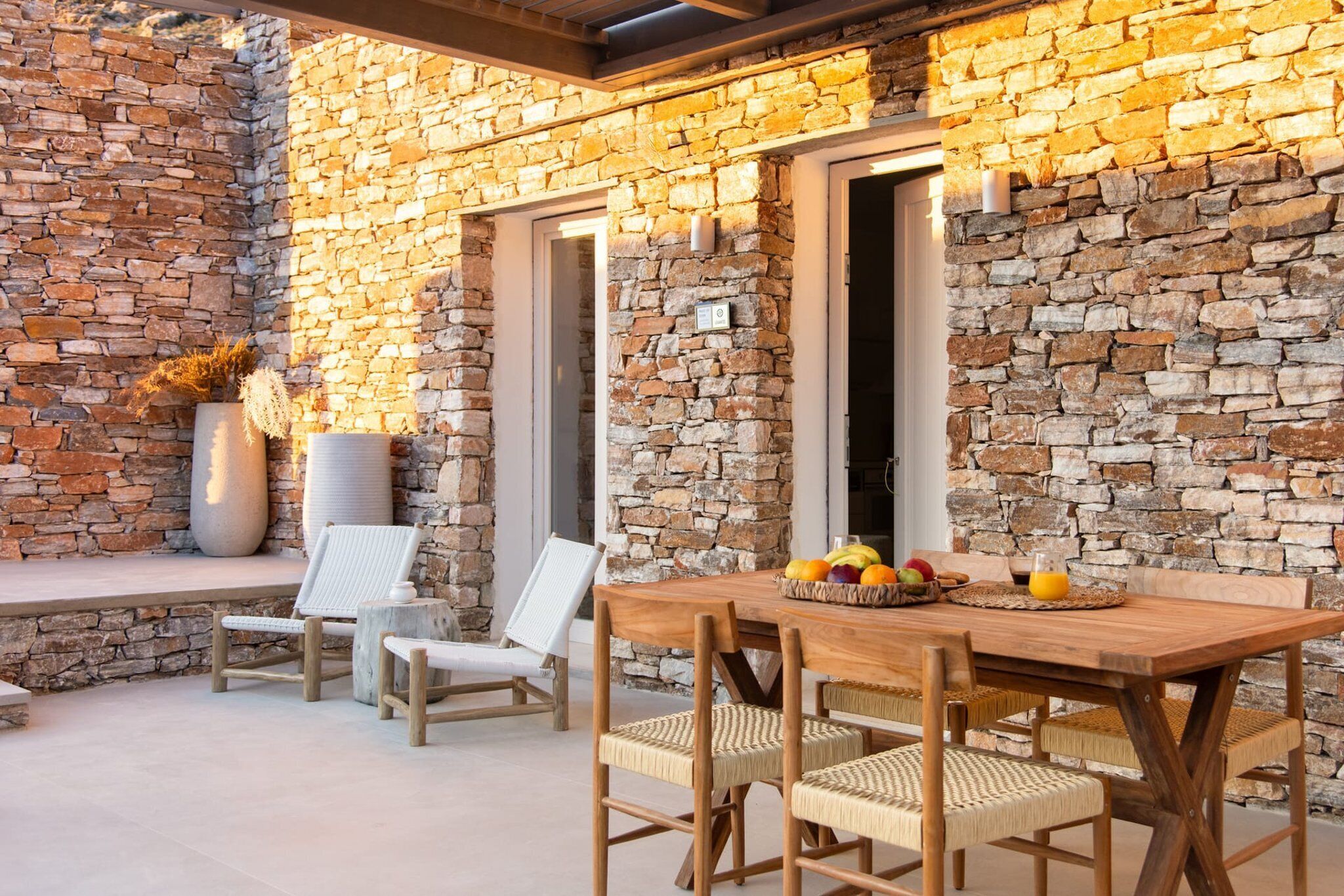 An outdoor patio with a stone wall, wooden table, wicker chairs, and a fruit basket.