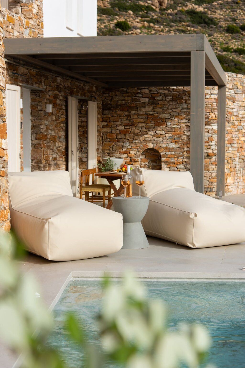 Ein Sitzbereich am Pool mit zwei beigen Sofas, einem kleinen runden Tisch mit Flasche und Glas sowie einer Wand aus Stein im Hintergrund.