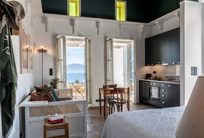 Una habitación con cama, zona de comedor con mesa y sillas, cocina con armarios oscuros y ventanas amplias que dan al mar.