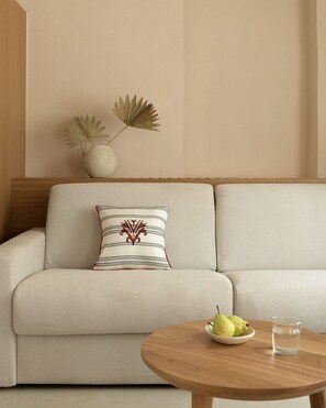A beige sofa with a decorative pillow, a wooden side table with a bowl of fruit and a glass of water, and a vase with two plants on the wall. - Majestic Rethymno Villa | Villa Helios Retreat | 3-Bedroom Luxury Residence |. (Rethymno)
