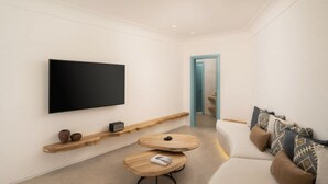A modern living room with a flat-screen TV mounted on the wall, a wooden shelf with decorative items, a round coffee table, and a sofa with patterned cushions. - Lavish Santorini Suite | Sunset Embrace Suite | Executive Two Bedroom Suite. (Santorini)