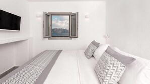 A bedroom with a bed, a TV mounted on the wall, a window with a view of the sea, and a patterned rug. - Thrilling Santorini Room | Poseidon’s Hideaway Room | Breathtaking Caldera and. (Oia)