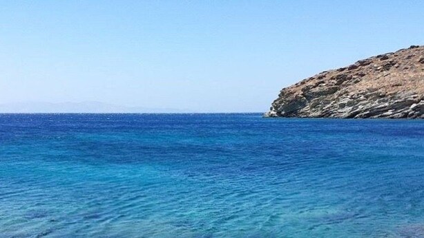 Zwei Holzliegen, die auf das klare, blaue Meer zeigen, mit einer felsigen Landzunge im Hintergrund.