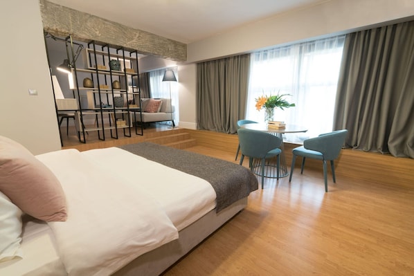 A modern hotel room with a large bed, a seating area with two chairs, a small table, and a bookshelf.