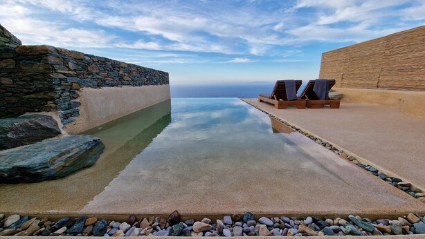 Una piscina infinita con un muro de piedra en un lado y una terraza de madera con sillones de descanso en el otro.