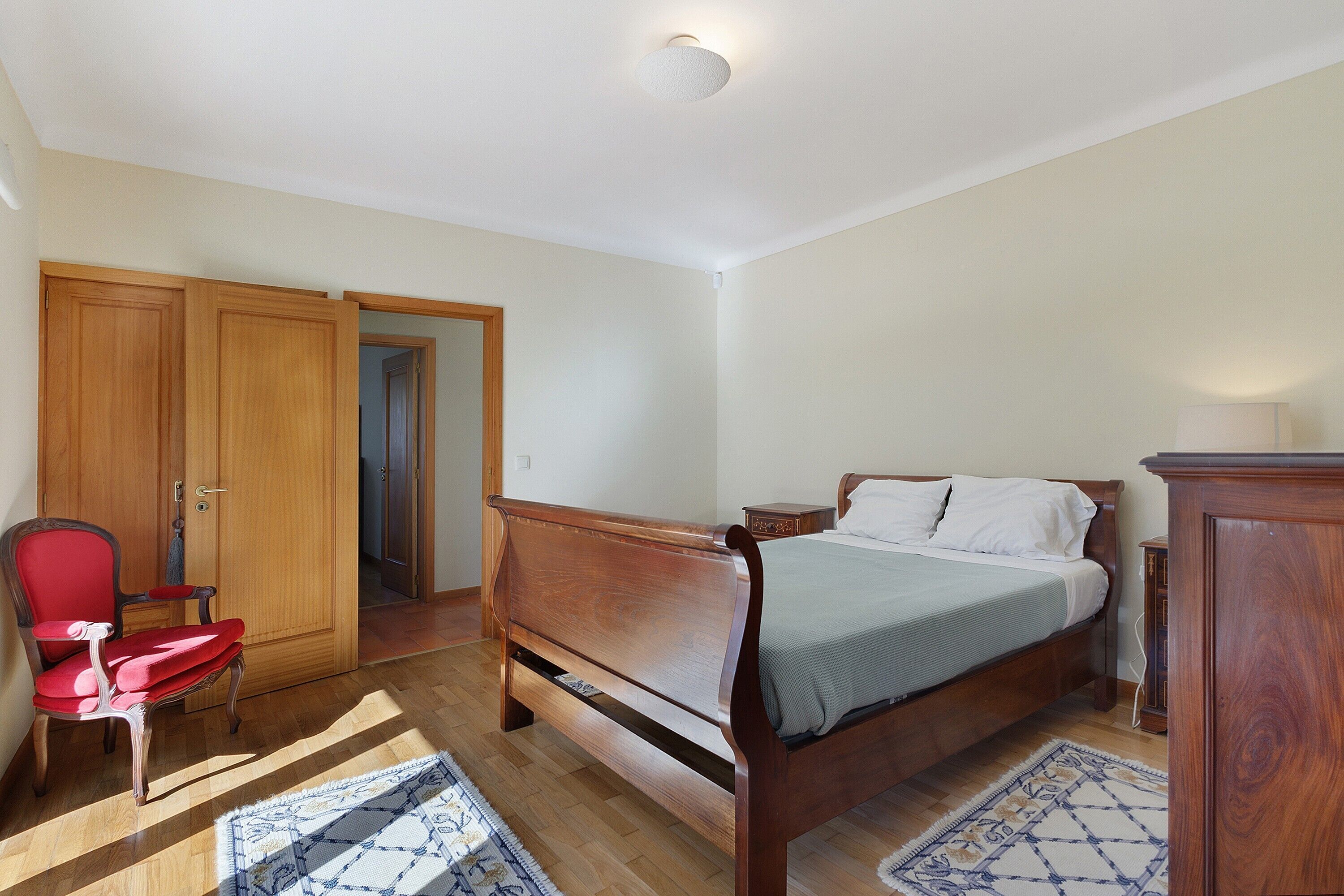 A bedroom with a wooden bed, a red chair, a wooden wardrobe, and a door leading to another room.