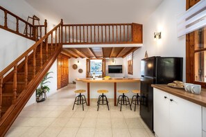 Eine Küche mit einer Holztreppe, einer zentralen Kochinsel mit Hockern, einem schwarzen Kühlschrank und Blick ins Freie.