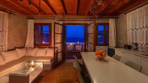 Ett mysigt rum med ett träpanelat tak, en soffa, ett matbord med en fruktskål och utsikt över havet genom öppna fönster.