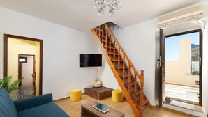 Ein Wohnzimmer mit blauem Sofa, Couchtisch aus Holz, gelben Hocker, einer Holztreppe und einem an der Wand befestigten Fernseher.