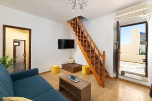 Ein Wohnzimmer mit blauem Sofa, Couchtisch aus Holz, gelben Hocker, einer Holztreppe und einem an der Wand befestigten Fernseher.