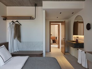 Ett modernt hotellrum med en säng, en bänk, en klädstång och ett badrum med spegel och handfat, samt en hall som leder till ett annat rum.