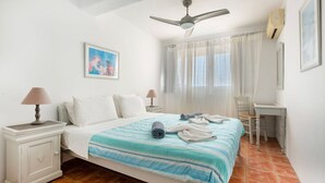 Ein Schlafzimmer mit einem Bett, einem Schreibtisch, einem Stuhl, einem Deckenventilator und einem Fenster mit Vorhängen.