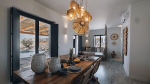 Un espace repas moderne avec une table en bois prévue pour quatre personnes, de grandes suspensions en fibres tressées et une vue sur une piscine et un espace extérieur grâce à de grandes fenêtres.