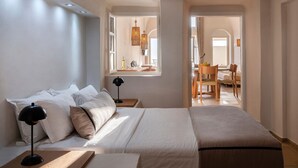 A modern hotel room with a large bed, bedside tables, a window with a view, and a dining area visible through an open door. - Phoebeia Suite, Hot Tub & Caldera View, Imerovigli (Santorini)