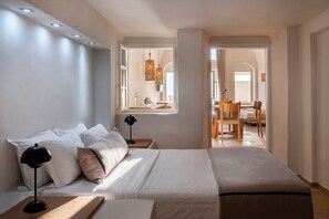 Une chambre d’hôtel moderne dotée d’un grand lit, de tables de chevet, d’une fenêtre offrant une vue agréable et d’un coin repas visible à travers une porte ouverte.