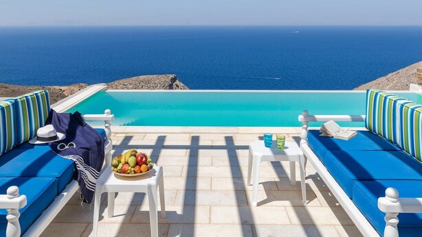 Ein Poolbereich mit Meerblick, zwei weiße Tische mit Stühlen und blau gepolsterte Sitzmöglichkeiten.