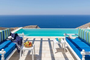 Area piscina con vista sul mare, due tavoli bianchi con sedie e comodi posti a sedere imbottiti di blu.